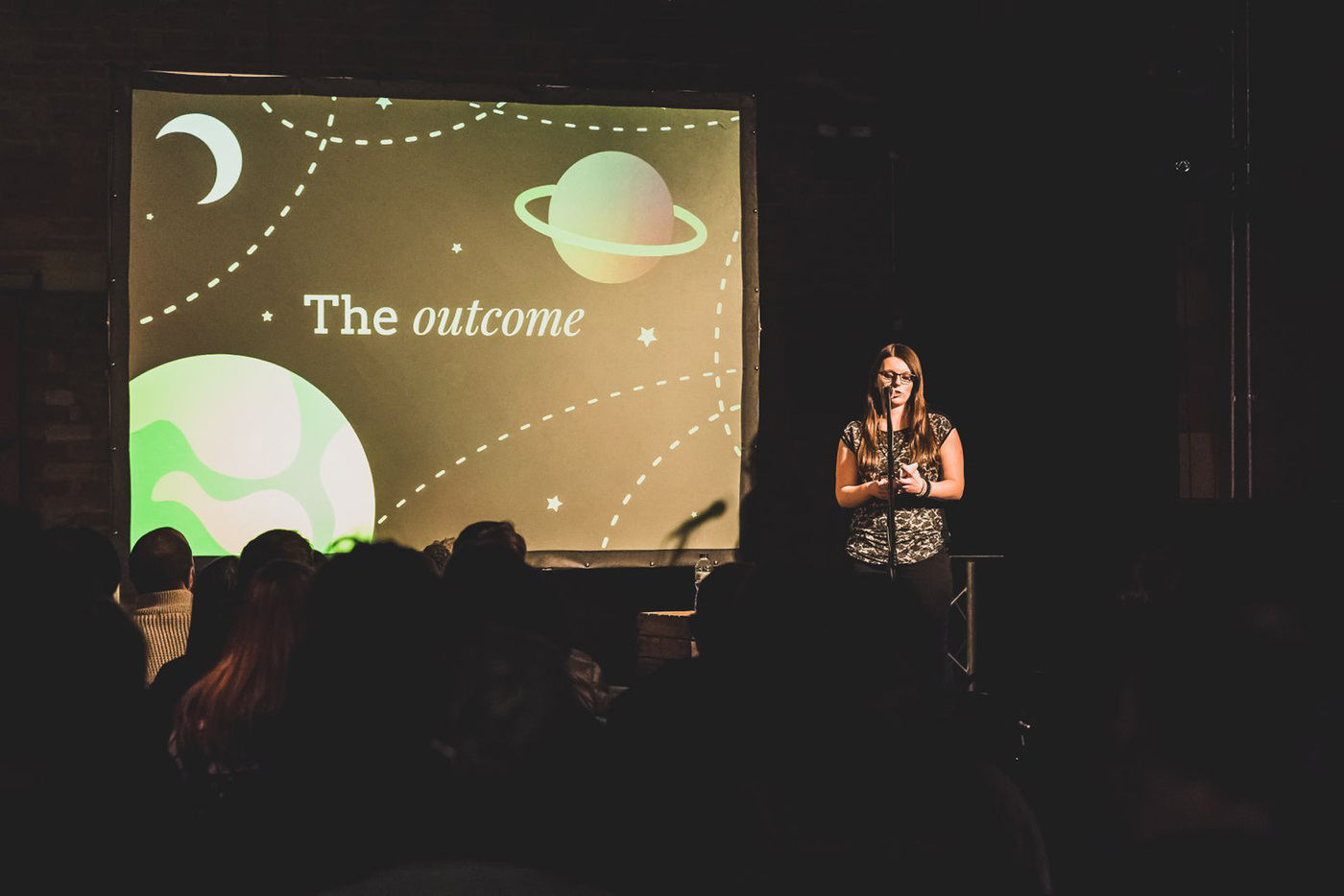 Christine on stage with a microphone and a projector screen on the left. The projector screen shows a slide saying 'The outcome', accompanied with illustrations of planets and stars. 